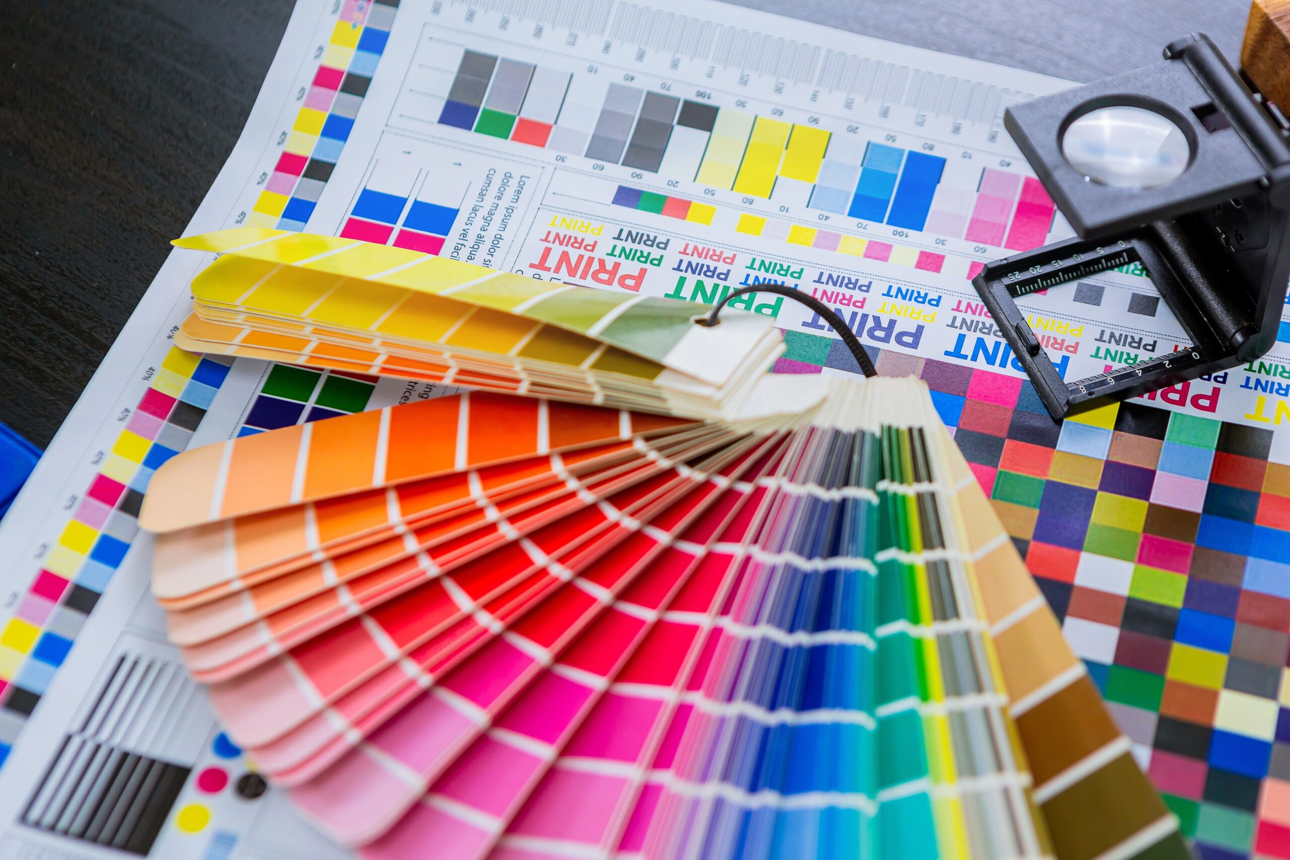 This high-angle shot captures the precision of the graphic arts industry, featuring a vibrant color swatch fan spread across a technical printer's proof. A professional linen tester magnifying glass sits atop the sheet, highlighting the focus on color accuracy and calibration. The composition showcases CMYK test bars and color grids essential for high-quality production.